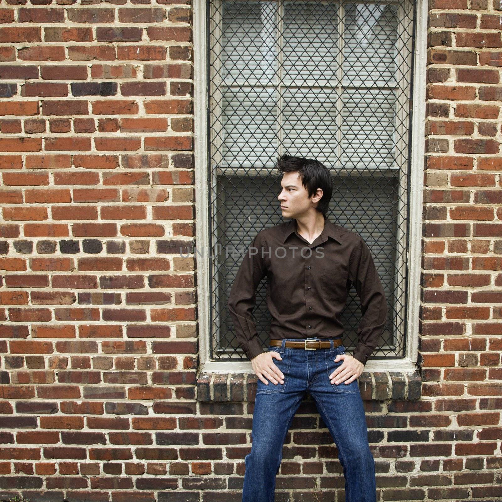 Asian man standing and leaning against window and brick wall.