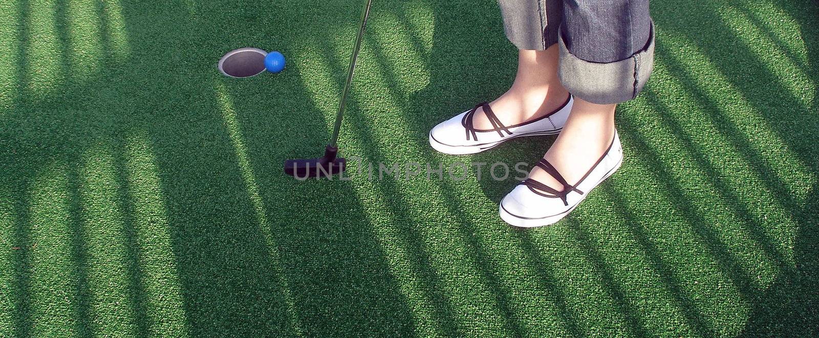 Playing adventure golf, feet, club, ball, hole and shadows.