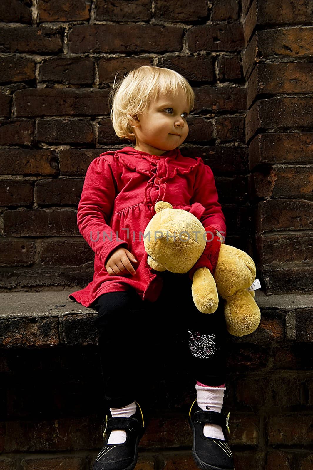 little blonde girl in red coat with teddy bear is seating on the window sill