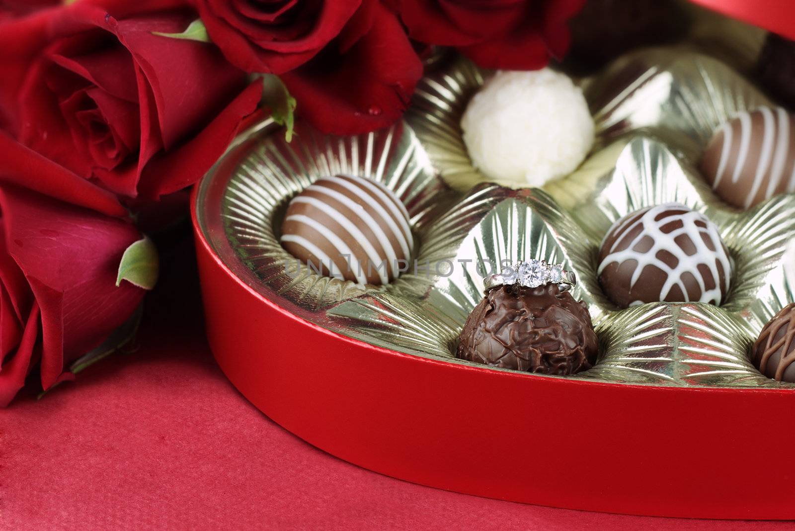 Diamond engagement inside of a heart shaped box of chocolate truffles with red roses. Selective focus on diamond ring with soft blurred background.