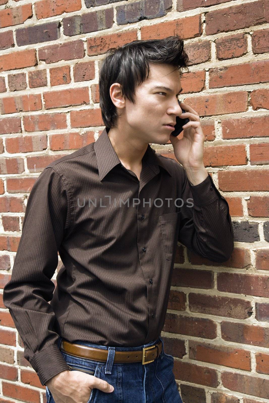 Young Asian man next to brick wall talking on cell phone.