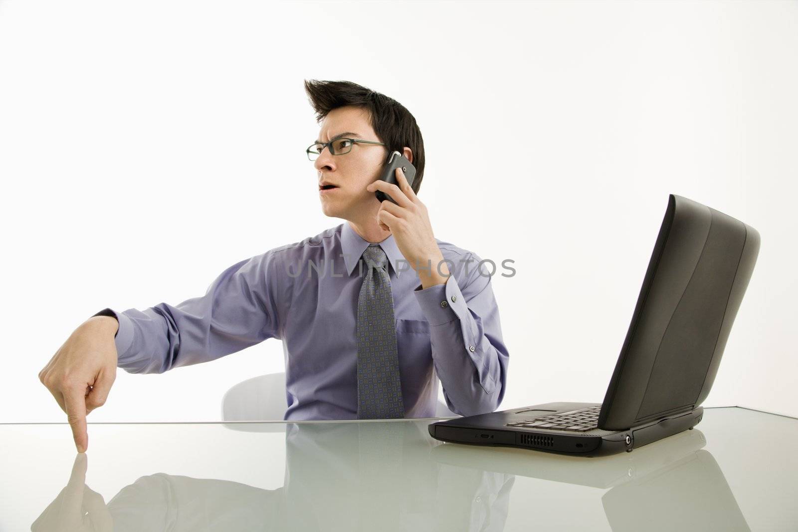 Asian businessman sitting at desk on cellphone with angry expression.