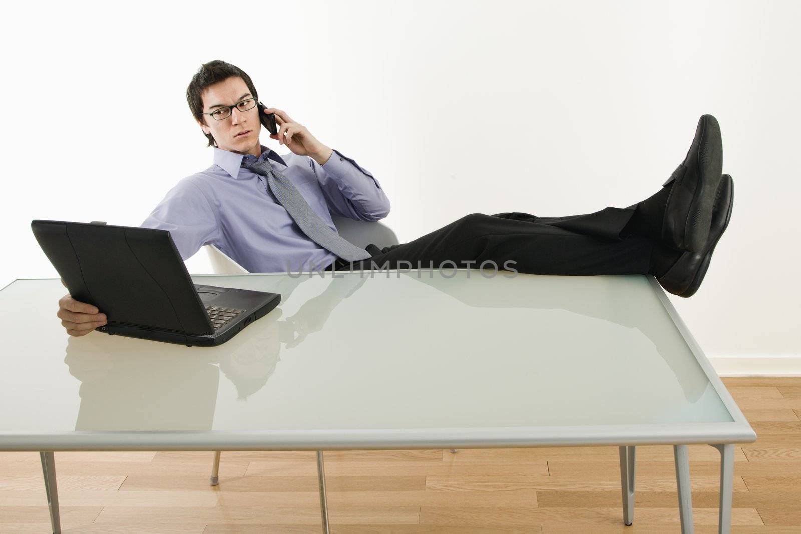 Asian businessman sitting at desk talking on cellphone and using laptop.