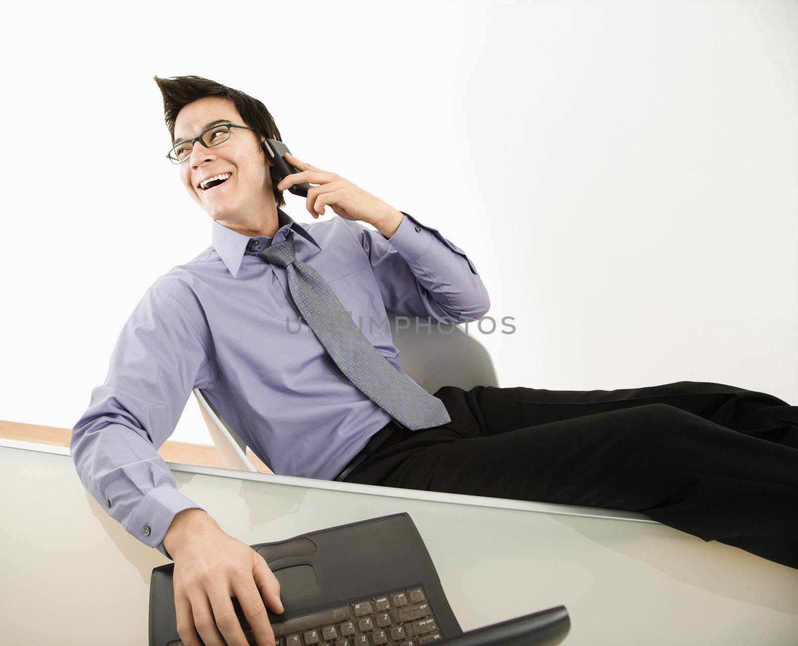 Laughing Asian businessman sitting at desk talking on cellphone.