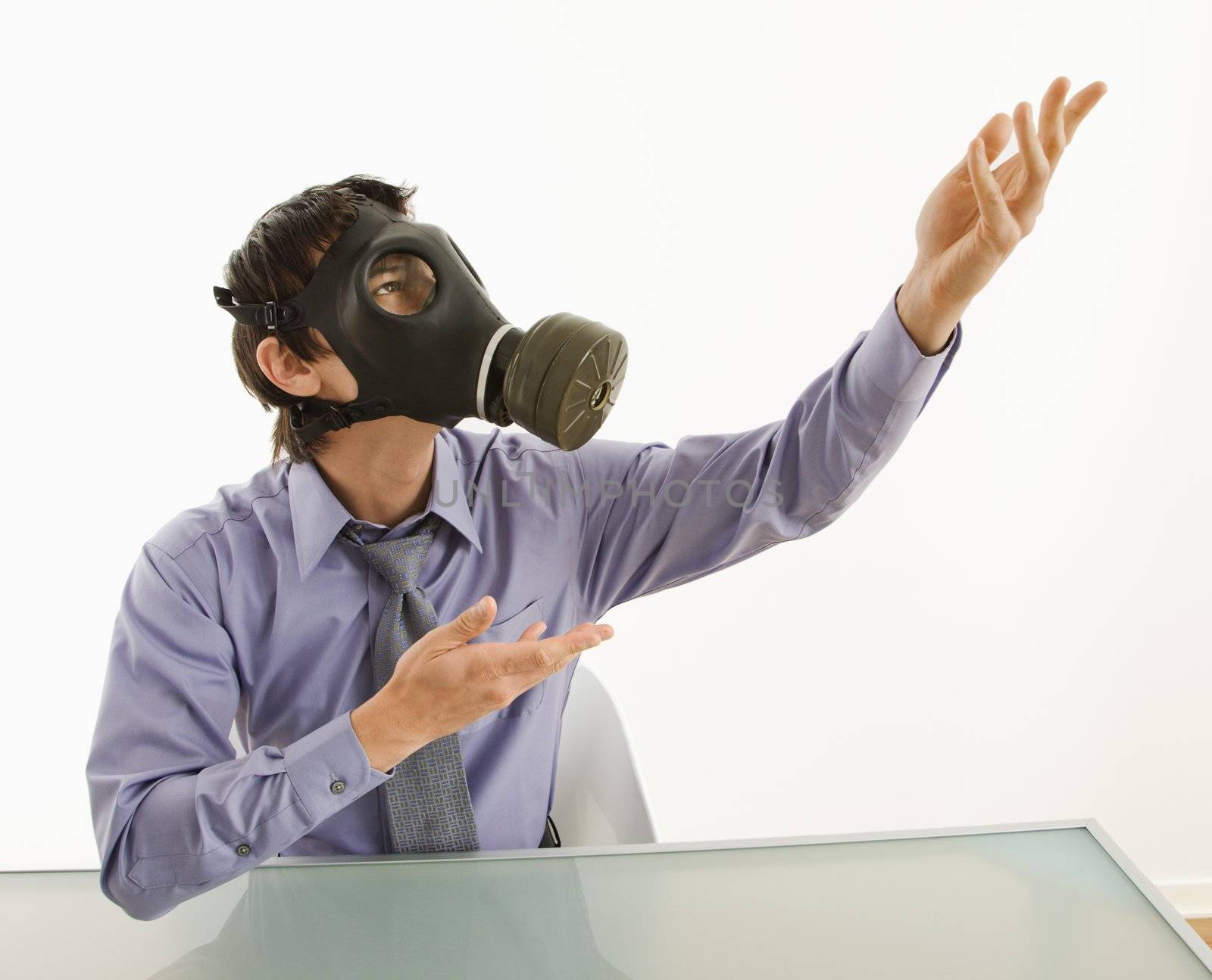 Businessman sitting in white room wearing gas mask gesturing to unknown person.