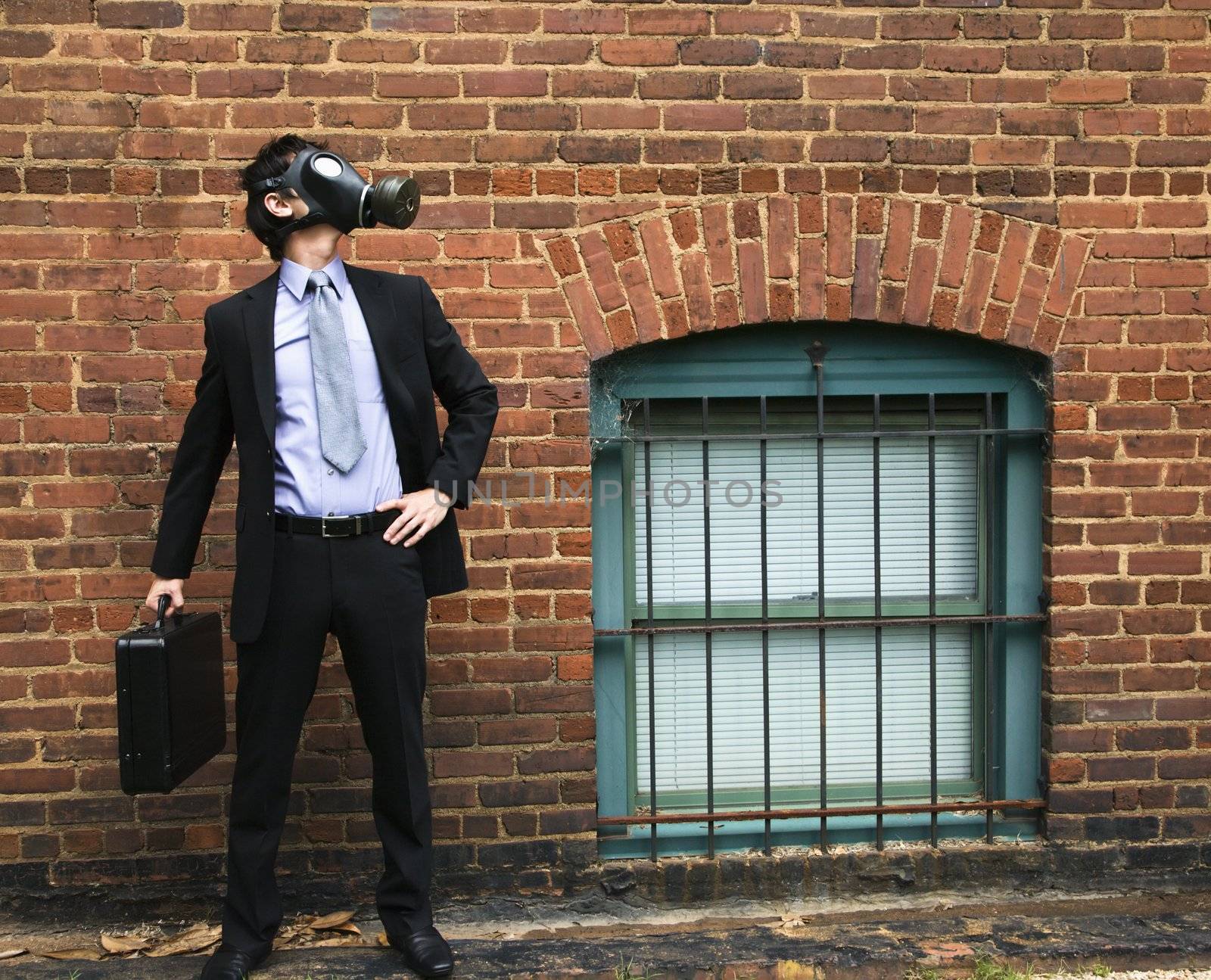 Businessman standing next to brick wall and looking off to side wearing gas mask.
