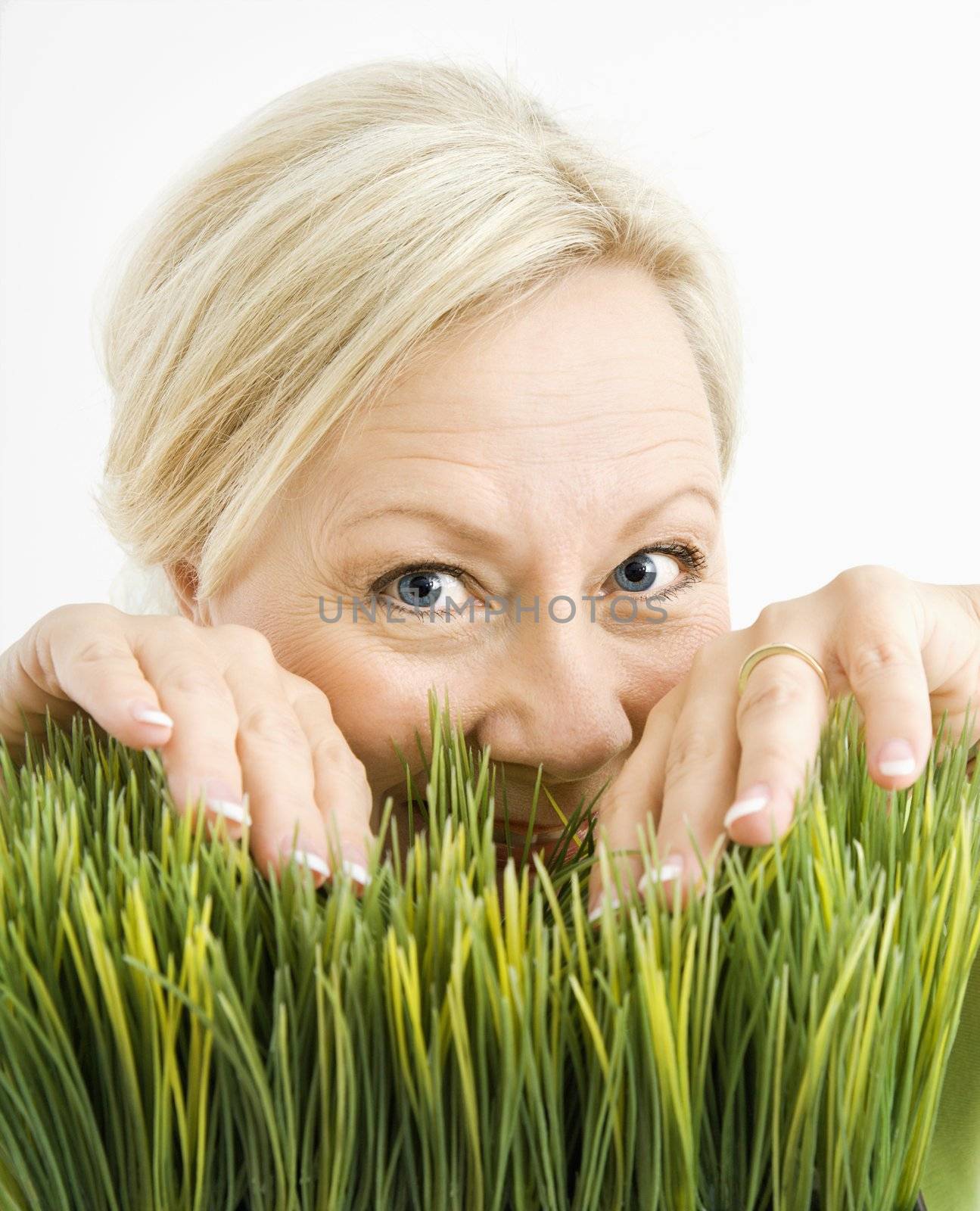 Portrait of adult blonde woman looking through grass at viewer.