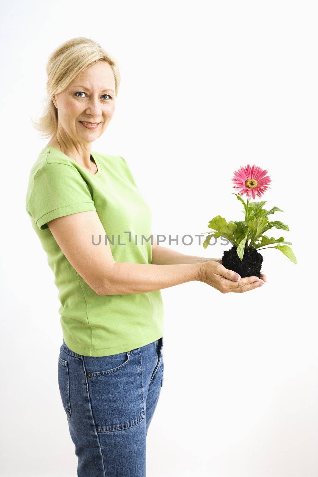 Side view of smiling adult blonde woman standing holding pink gerber daisy plant.