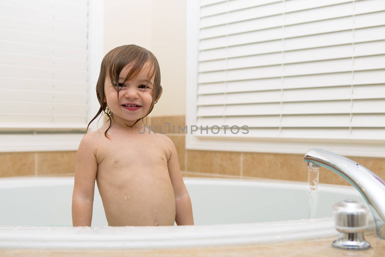 Toddler girl smiling trustfully from bath tub. Water coming from the bath tub faucet.