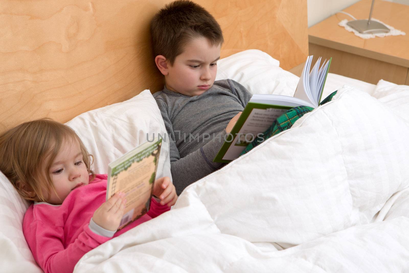 Reading habits starts with early age; siblings are reading their books before fall asleep. Perhaps cute toddler is just browsing the pages in her pink pajamas.