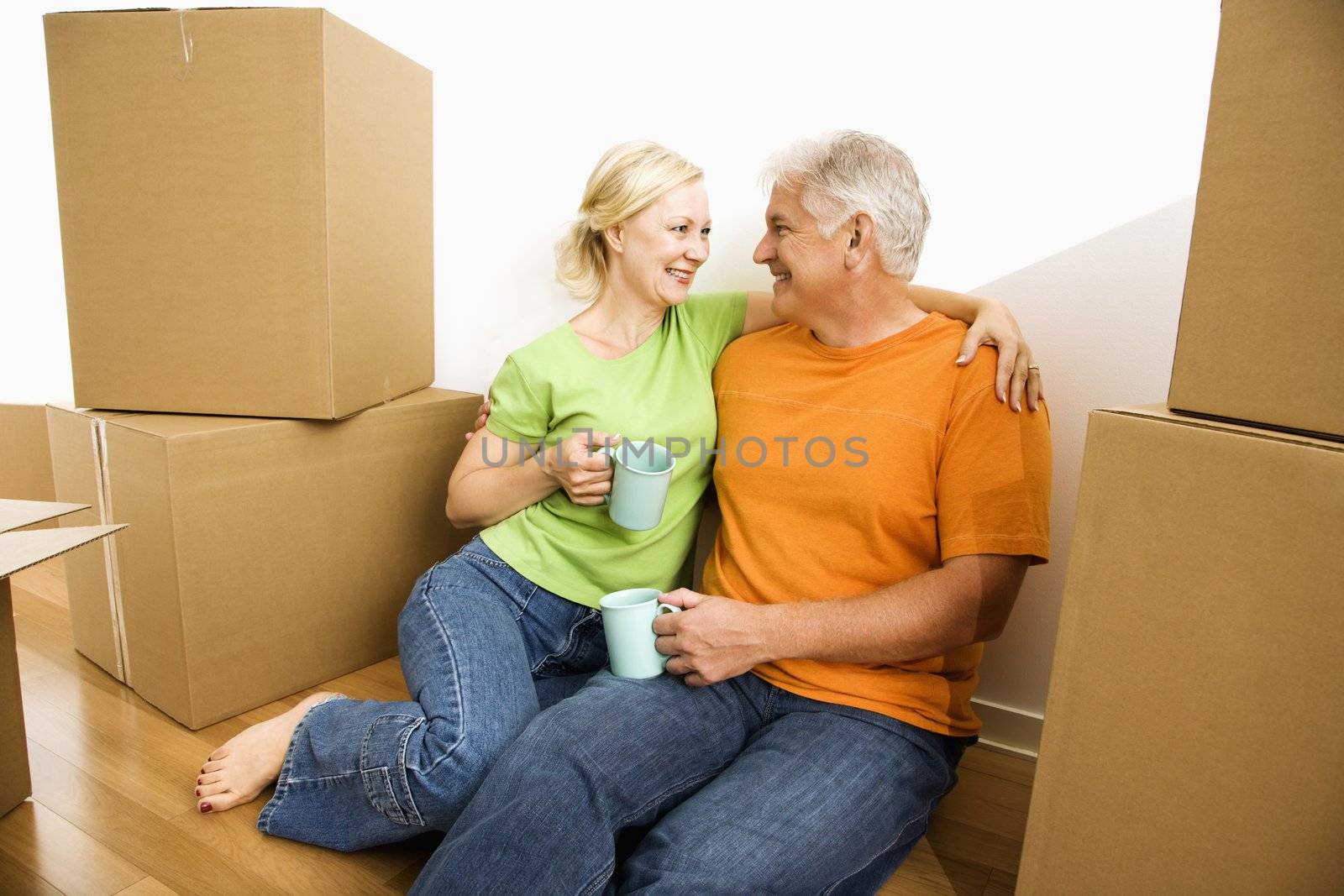 Middle-aged couple sitting on floor among cardboard moving boxes drinking coffee.