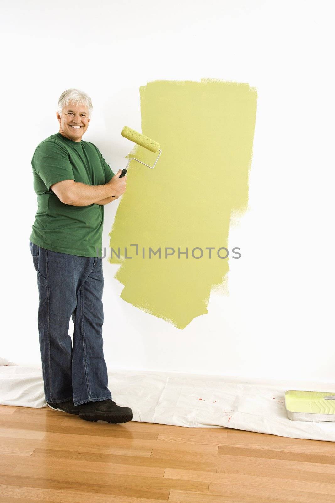 Middle-aged man painting wall green with paint roller smiling at viewer.
