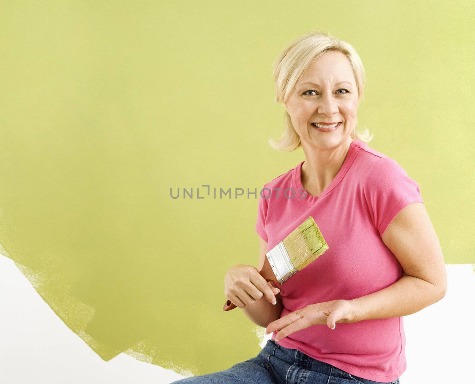 Portrait of smiling adult woman sitting in front of half-painted wall with paintbrush.