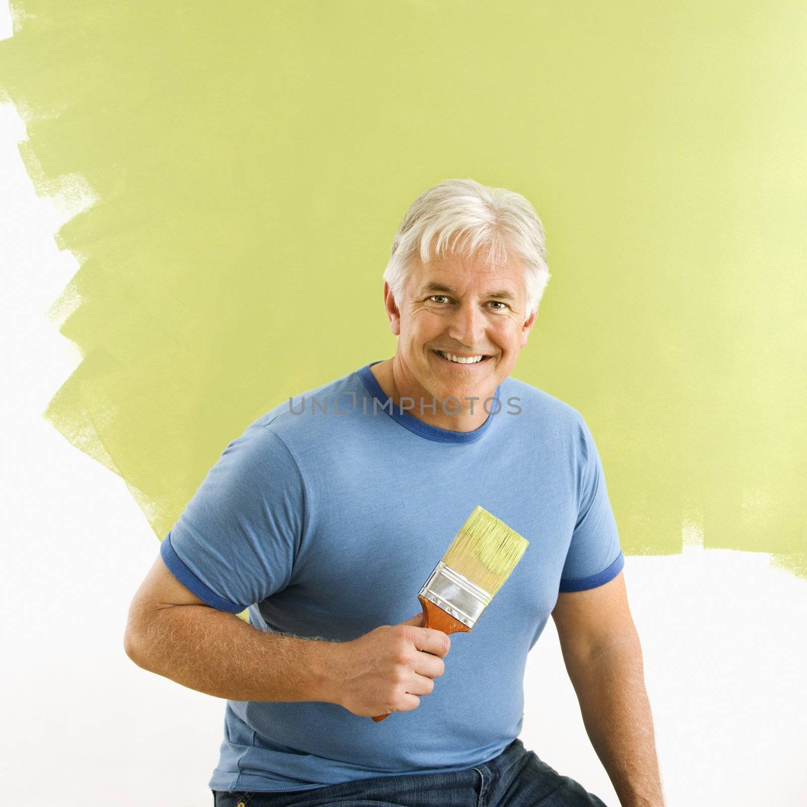 Portrait of smiling adult man sitting in front of half-painted wall with paintbrush.
