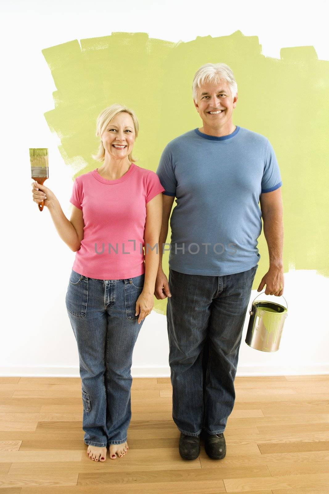 Portrait of happy adult couple standing in front of half-painted wall with paint supplies "American Gothic" style.