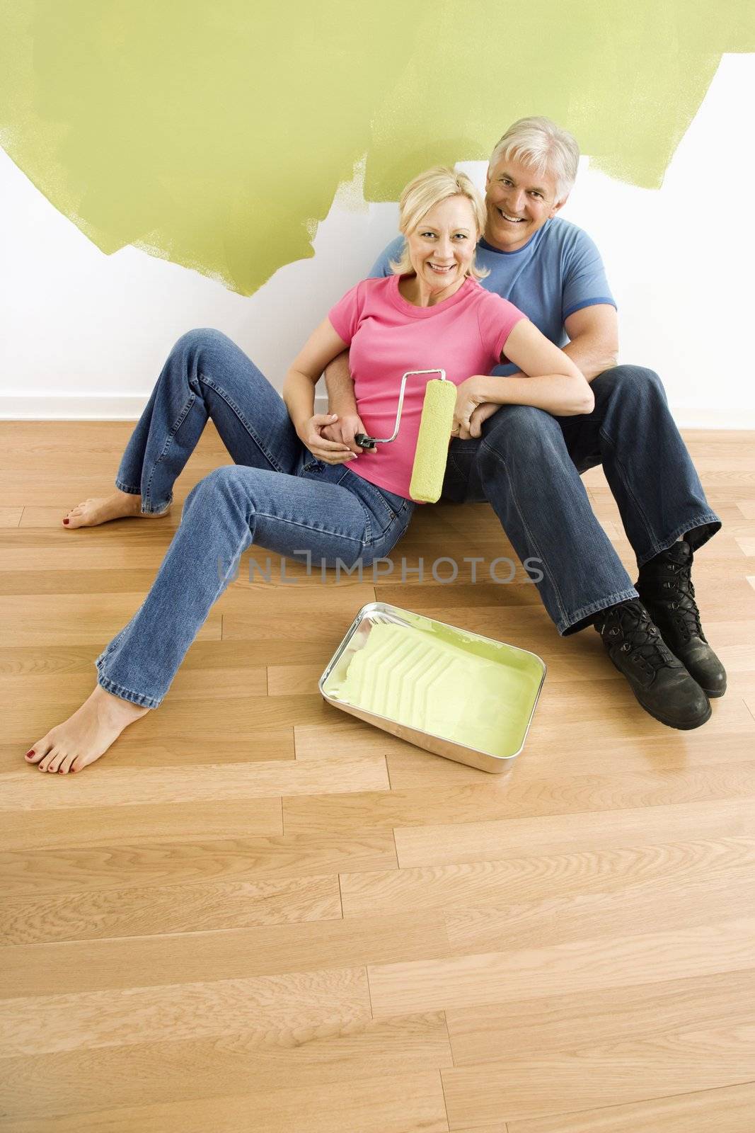 Portrait of happy adult couple sitting in front of half-painted wall with paint supplies snuggling.