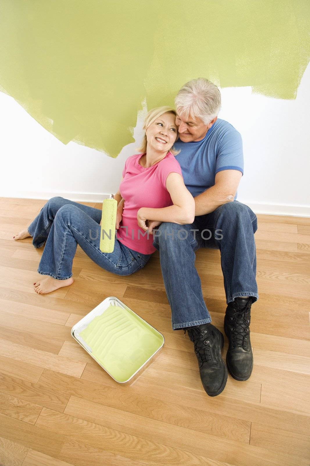 Portrait of happy adult couple sitting in front of half-painted wall with paint supplies snuggling.