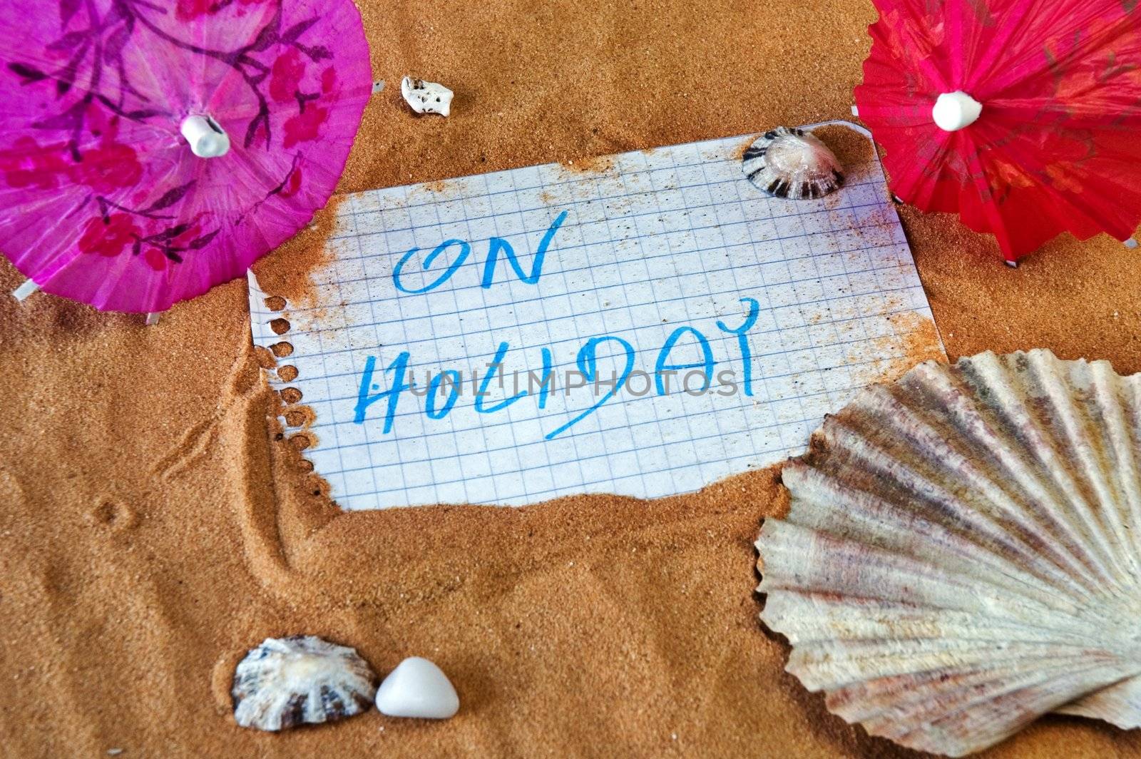 Notepad sheet on sand with On Holiday message with seashells and party umbrellas