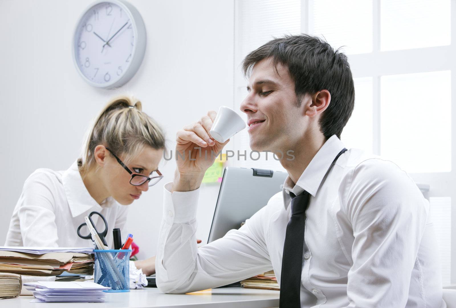 Business man enjoys a hot cup of coffee in the office, annoyed female colleague in the background