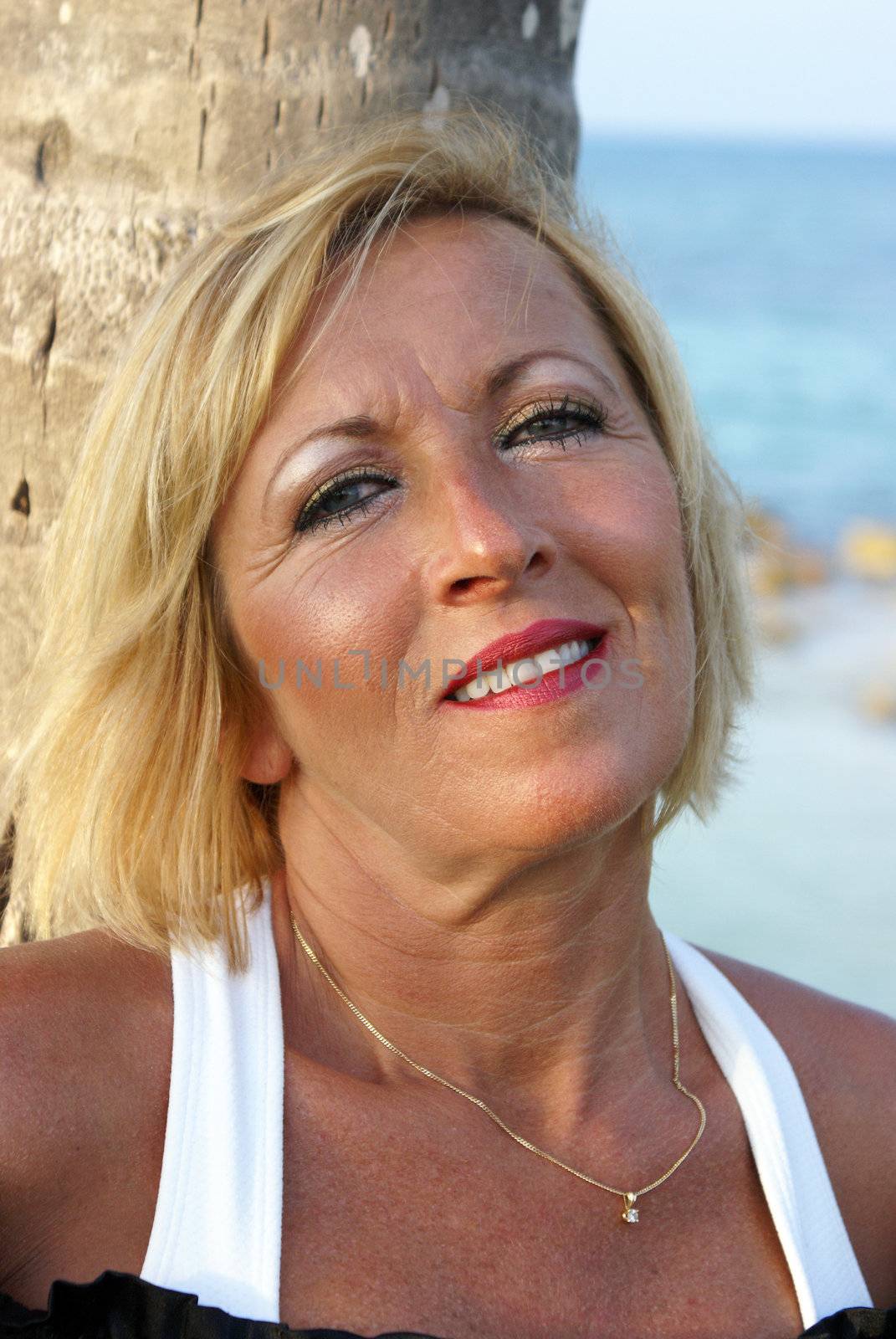 A woman in her 40's is looking at the viewer while leaning on a palm tree at the beach.