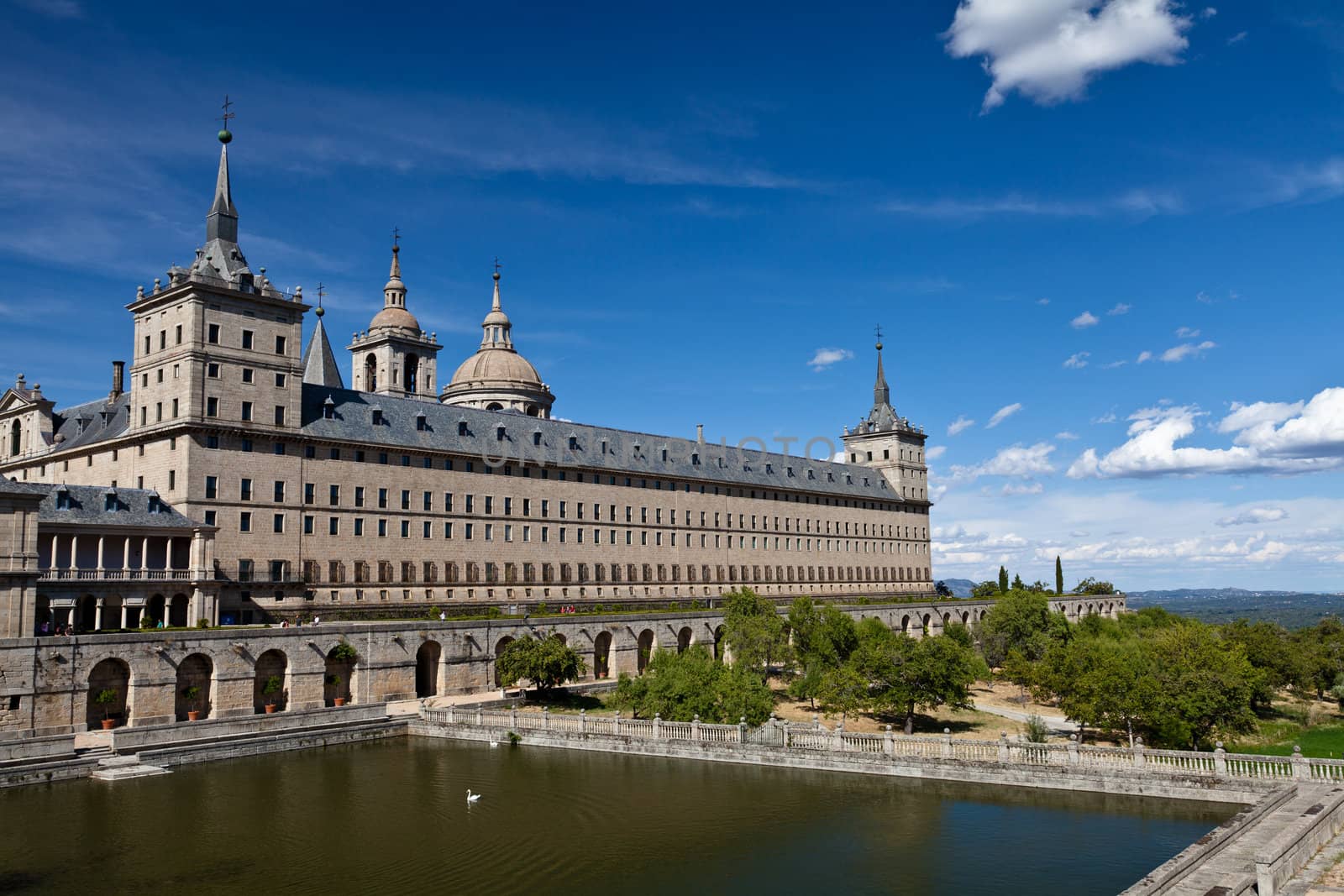 San Lorenzo de El Escorial Monastery with a reflecting pool with floating swan. The towers of the church and monastery are set of by a bright blue sky with a few white clouds.