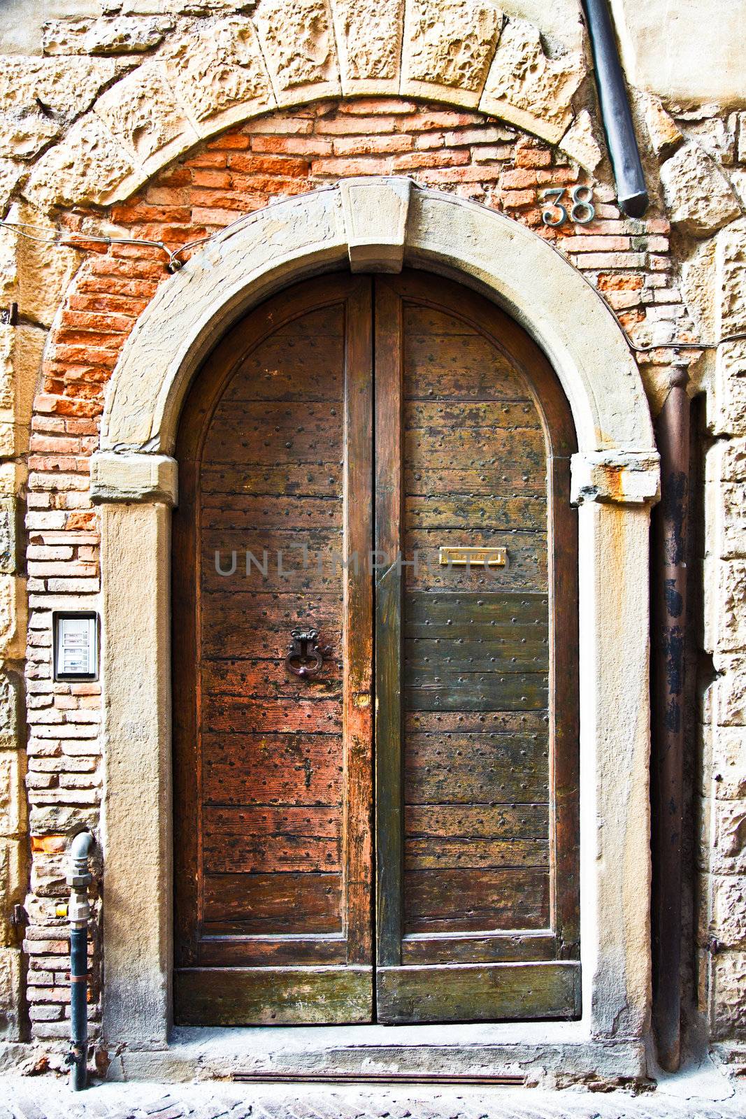 Old wooden door with a metal knocker and letter slot - wonderful texture. The door has little metal studs. On the side, there is a modern door bell and intercom. Exposed brick and pipes and a stone arch complete the picture.