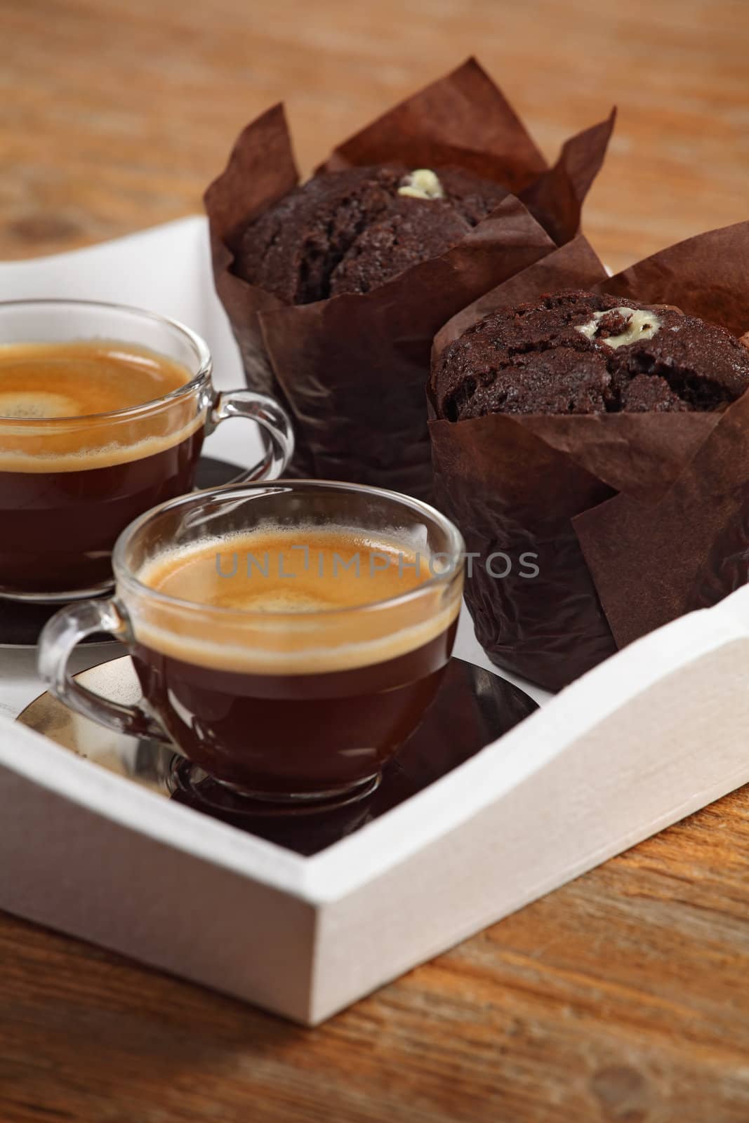 Photo of two moist chocolate muffins and two cups of espresso or coffee resting on a white serving tray. Shallow depth of field focusing across middle of photo.
