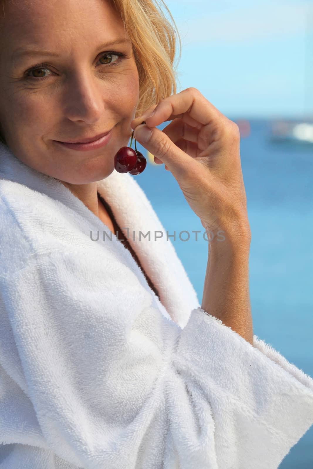 50 years old blonde woman dressed in bathrobe in front of the sea taking cherries in her fingers by phovoir