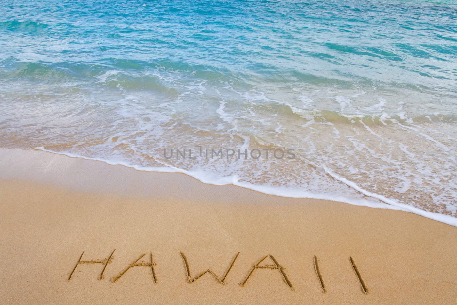 The word Hawaii is written in the sand of this beach as waves come in to wash it away. This is a vacation image showing the tropical location with sand and water of the Ocean.