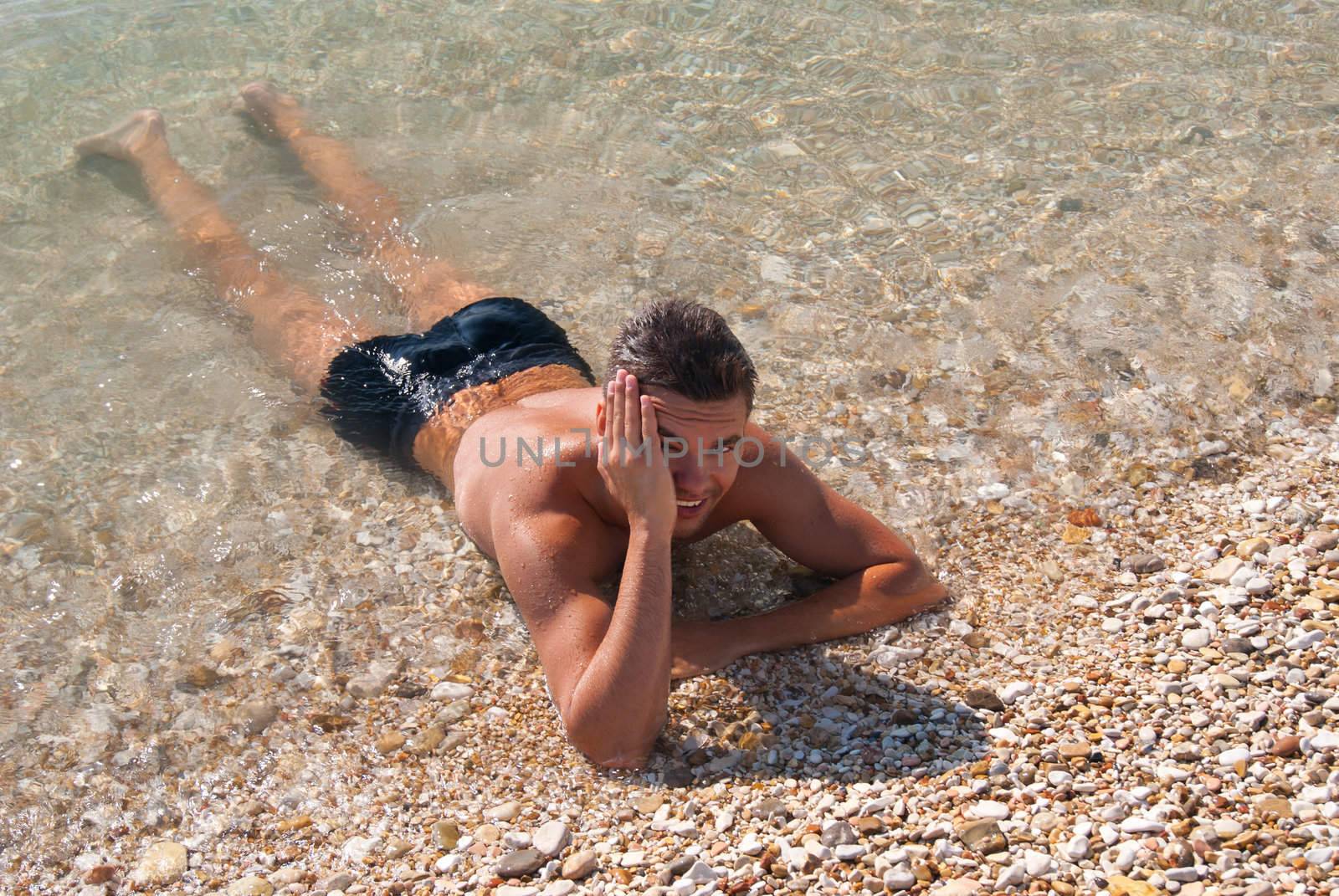 Beautiful tanned young fellow in sea water is shut by hand from sun in summer sunny day