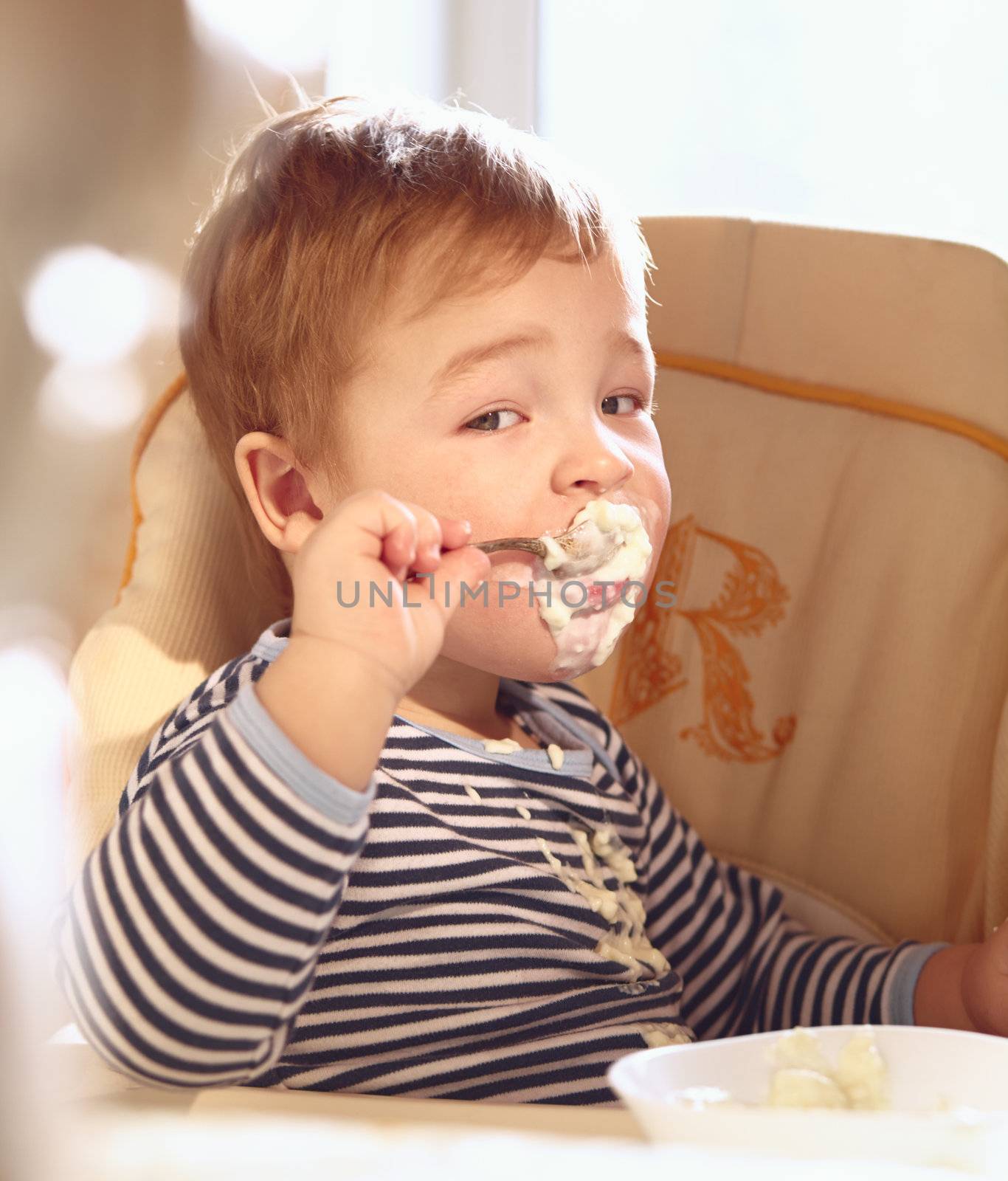 Portrait of two year old boy in the baby chair eating porridge in the morning.