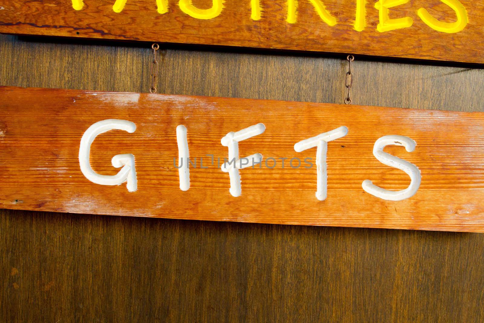 A handmade wood sign on a wood background is carved out and painted showing the word gifts in white letters.