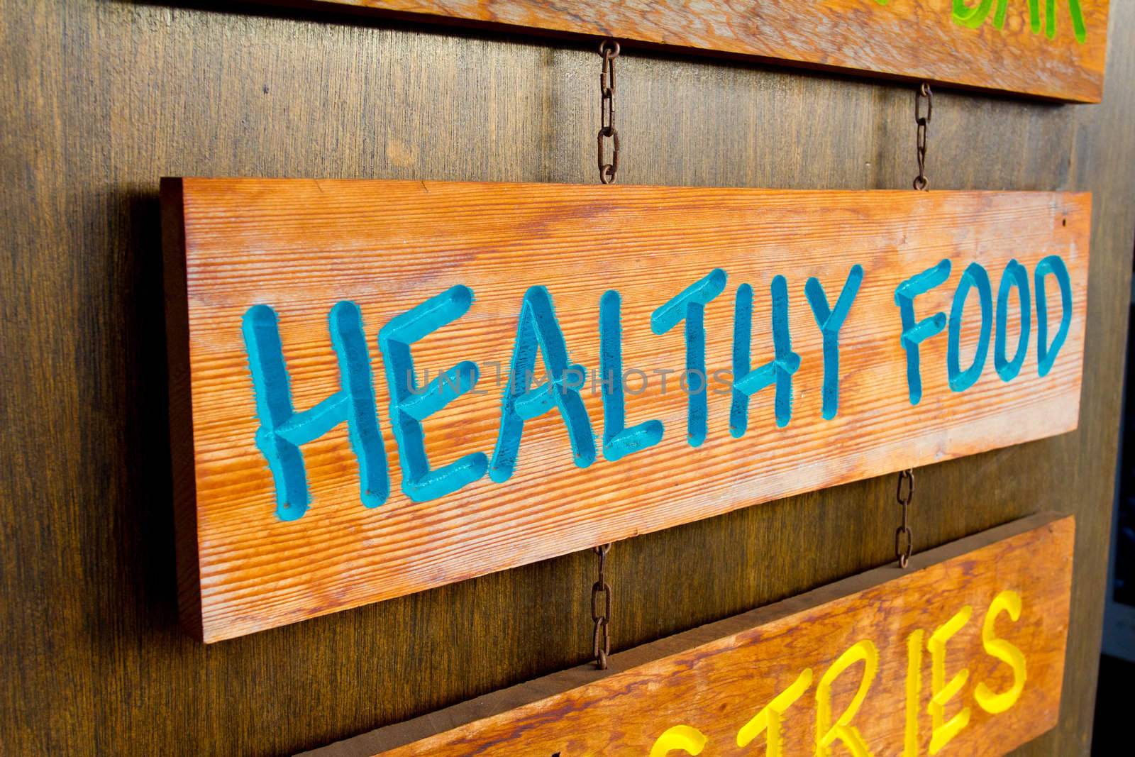A handmade wood sign on a wood background is carved out and painted showing the words healthy food in blue letters.