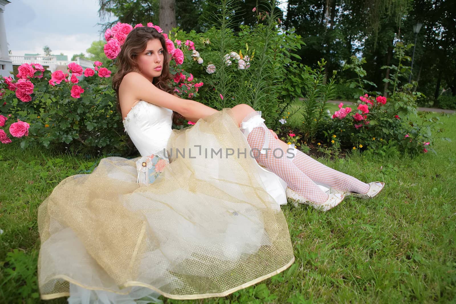 beautiful girl in wedding white-golden gown sits on herb in town park under rosebush