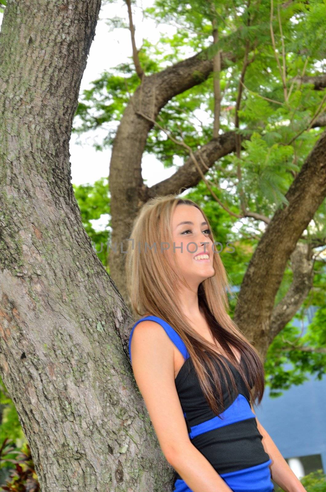 Attractive blond teenage girl lying back against a tree in a city park.