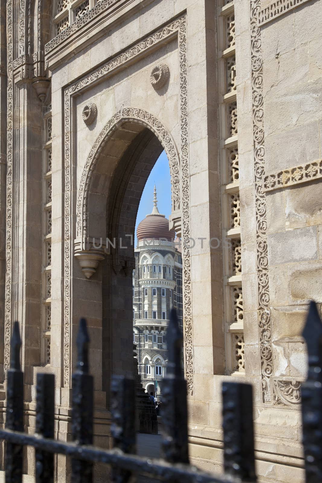 Vertical take of the Gateway of India, its black painted railings with a cameo view of the Taj Hotel from an angled view of an archway