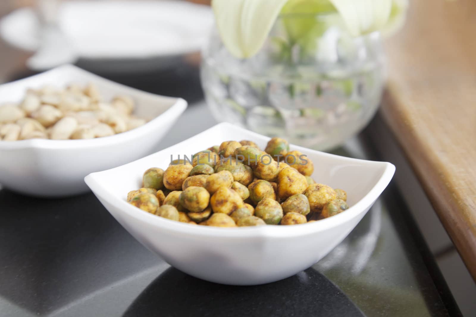 Horizontal studio setup of a bowl of dehydrated roasted spicy peas in a white square designer procelain square bowl on a table top with peanut bowl in background