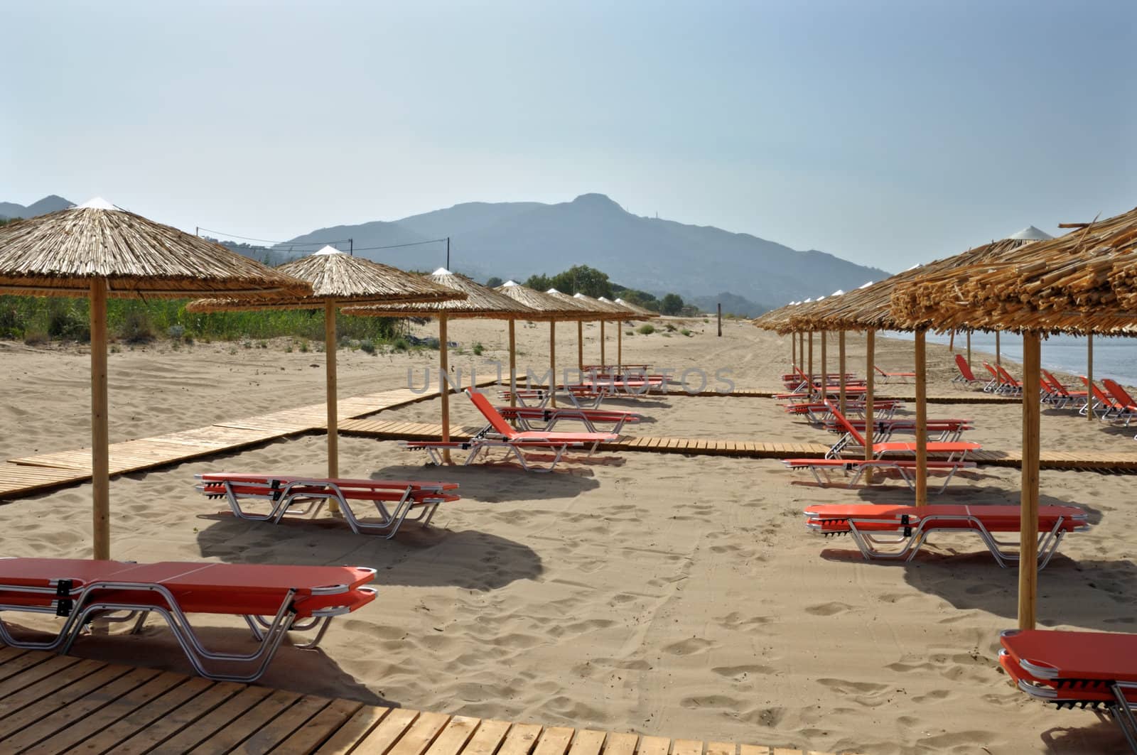 Straw umbrellas and rows of lounge sunbed chairs at sandy beach in Greece.