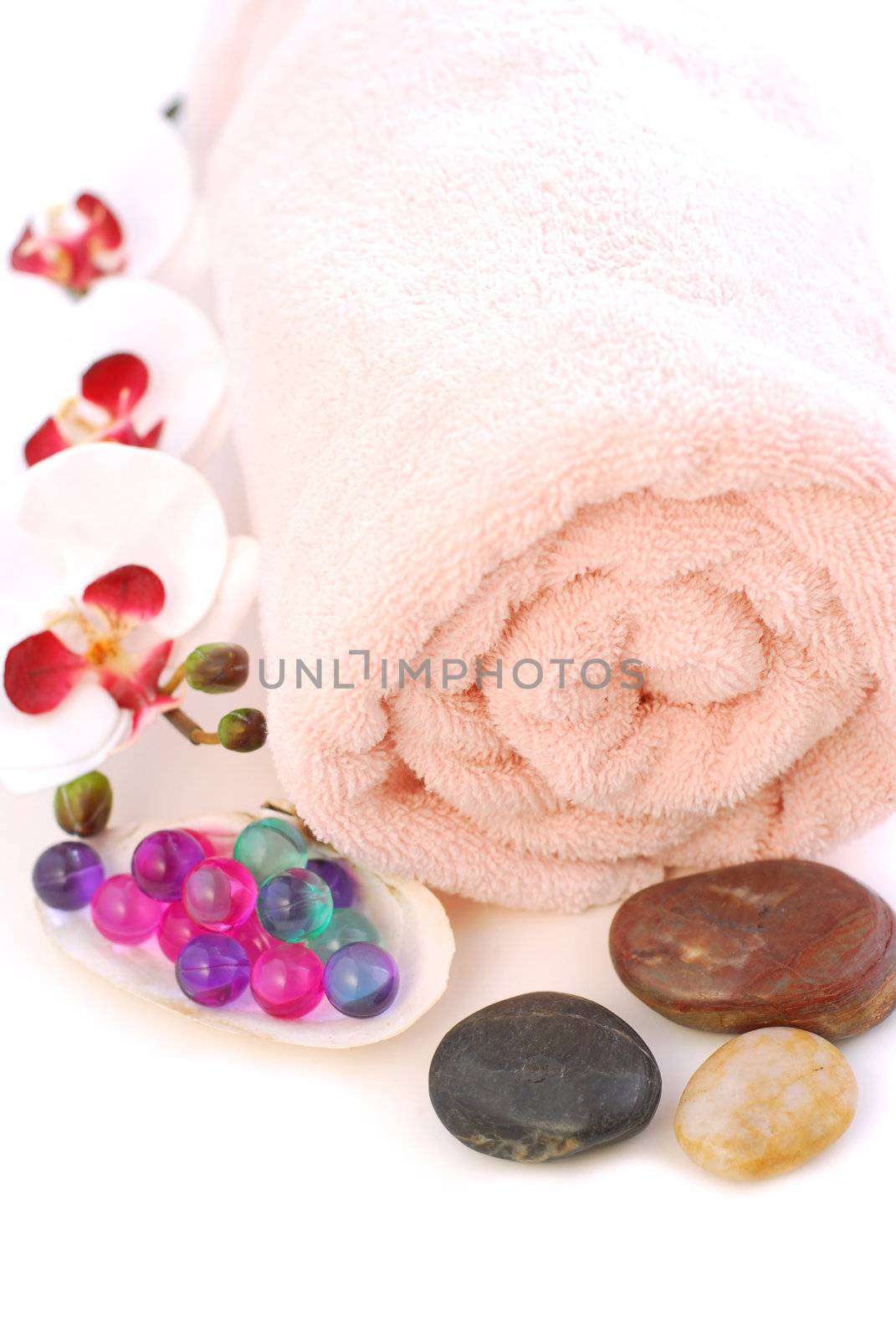 Pink rolled up towel with massage stones and bath beads on white background