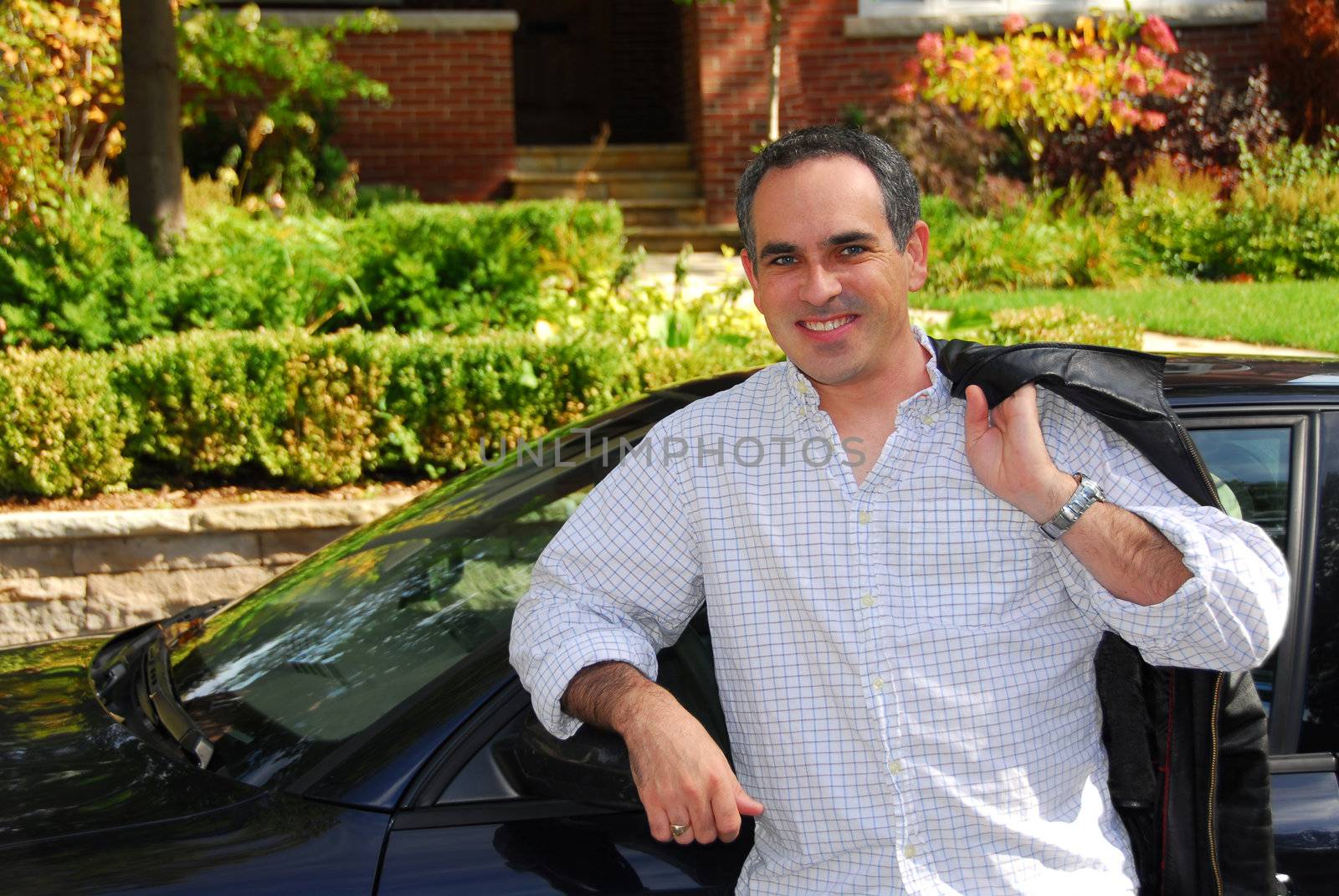 A man standing outside leaning on his car in front of his house