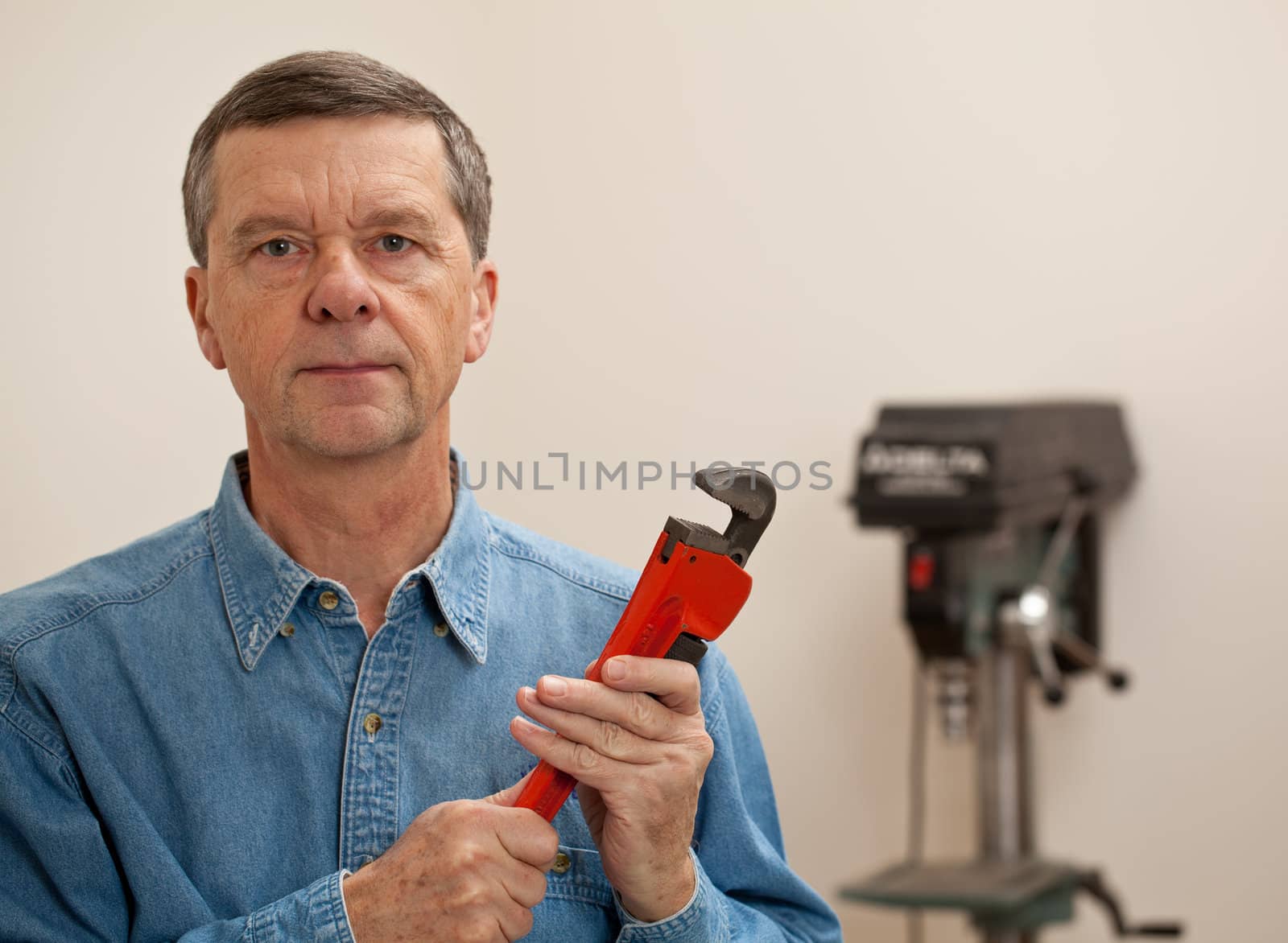 Senior male in a home workshop facing the camera and holding a large wrench