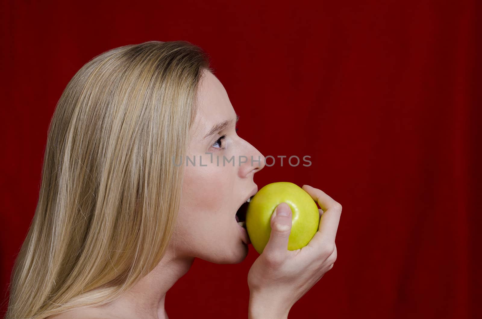 young blonde woman taking a bite of a green apple on red background