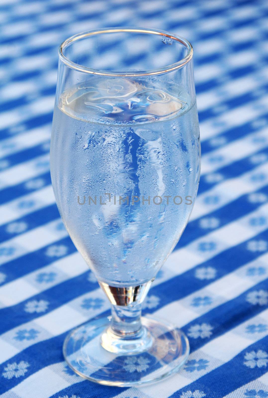 A glass of iced water, standing on a blue and white tablecloth, ice cubes in glass and condensed water on glass due to heat