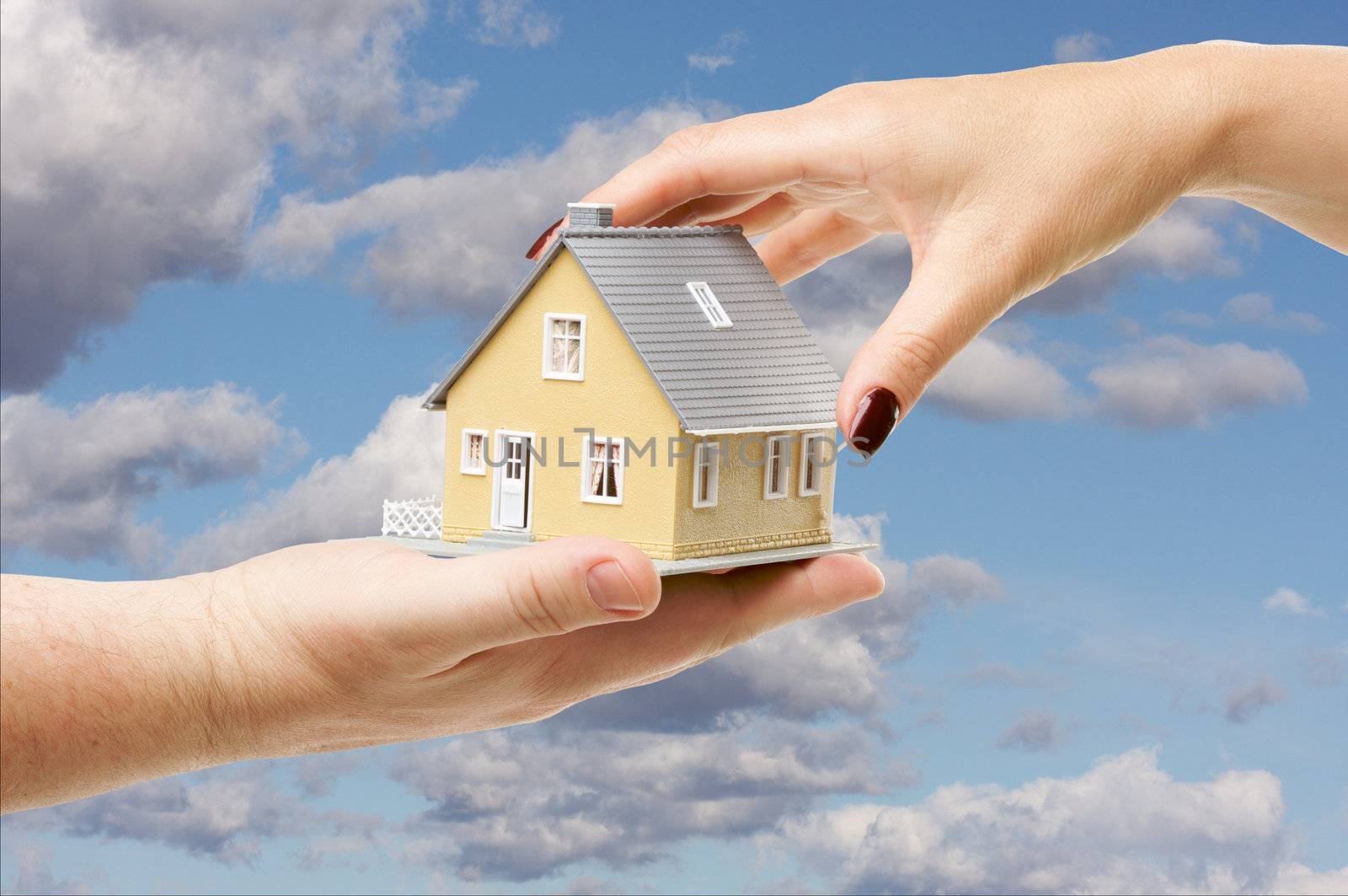 Female hand reaching for a house on a partly cloudy sky background.