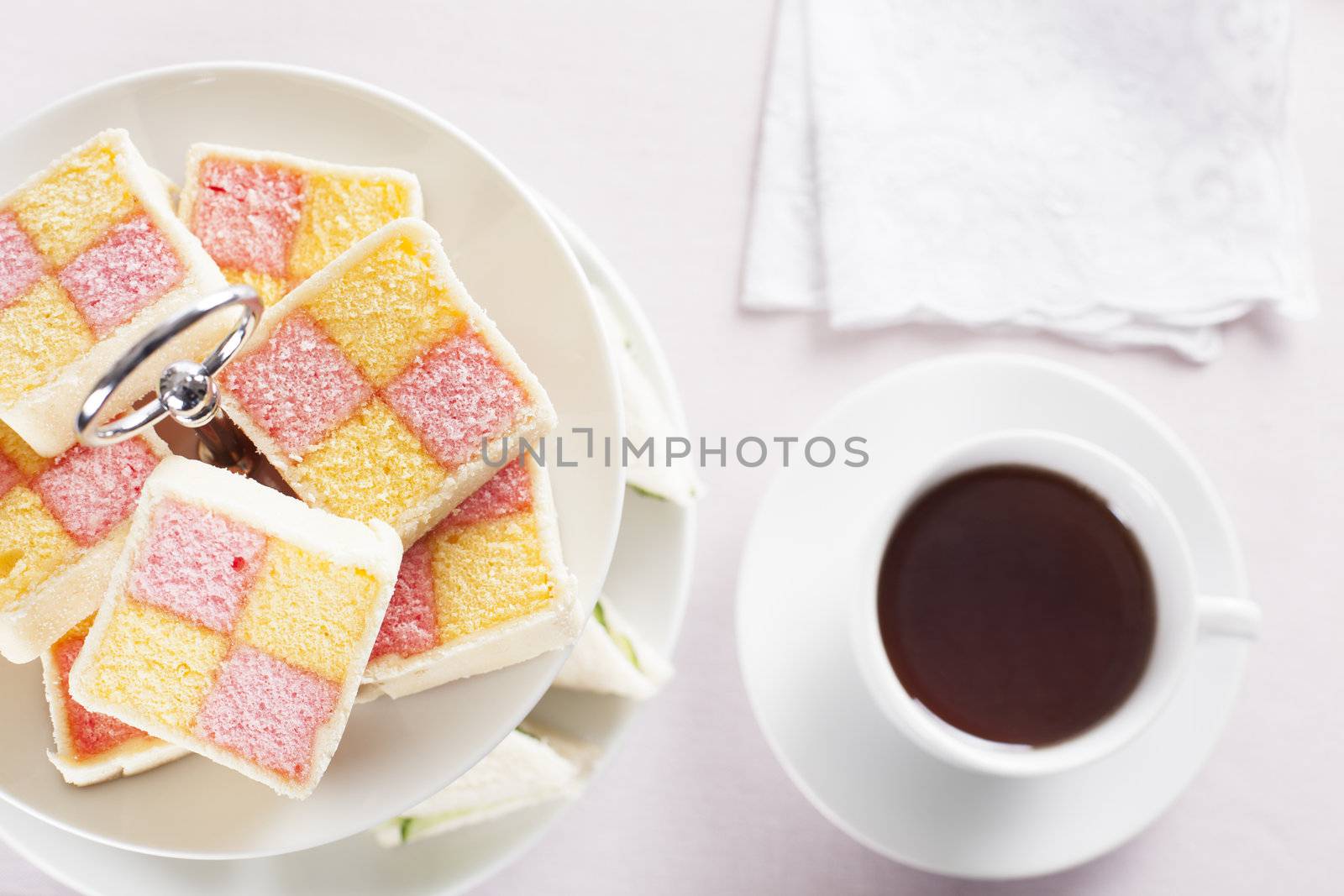 Battenberg cake on a two tiered serving plate and a cup of tea on the table