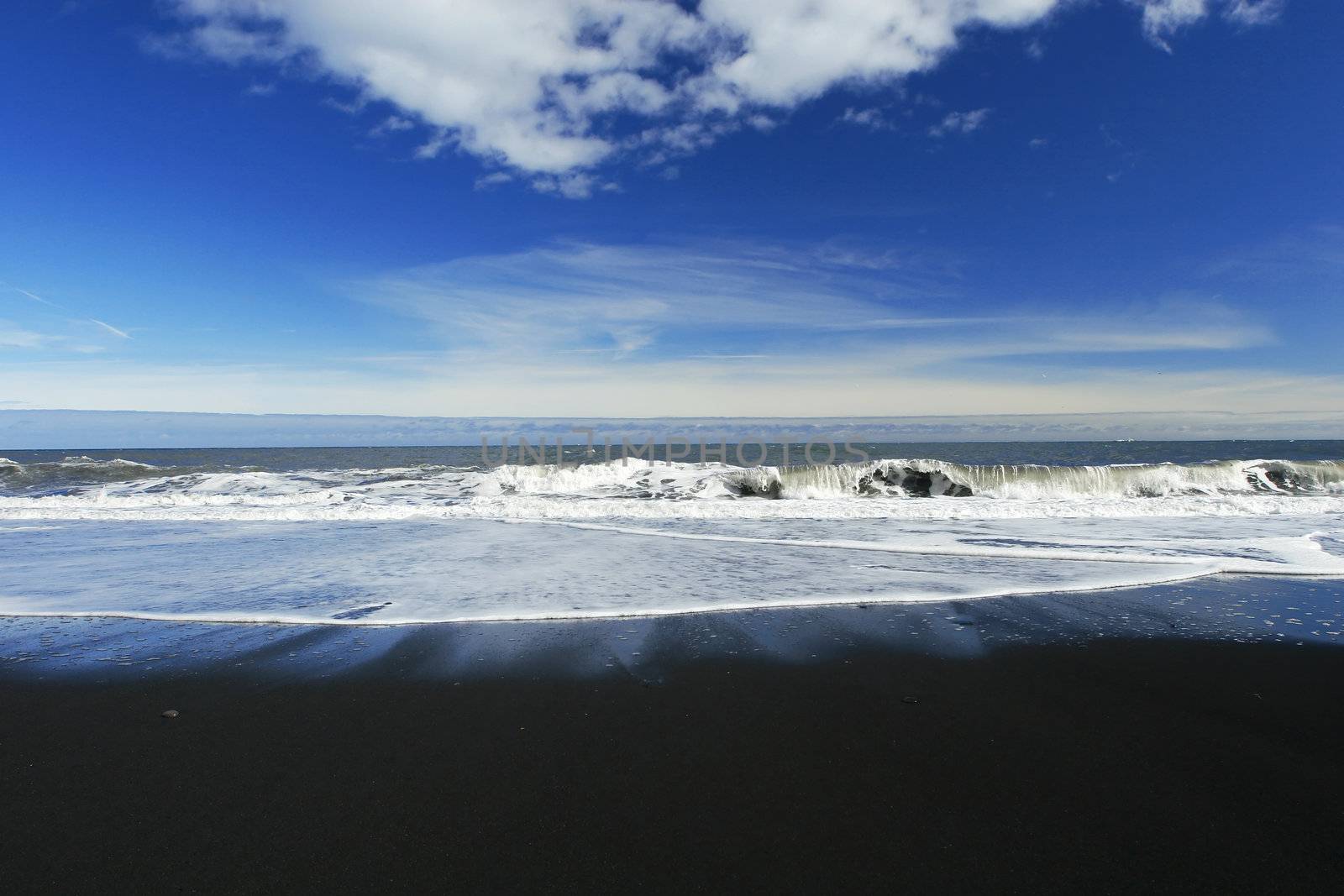 waves crashing on a sandy black beach , deep blue sky and clouds in background