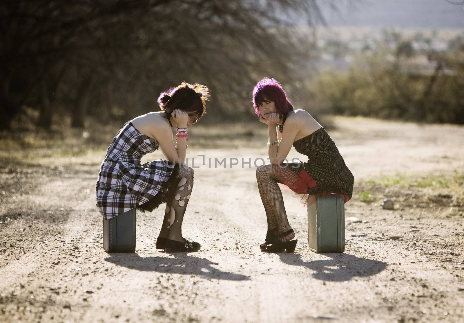 Two punk women sitting on suitcases and waiting on a rural road