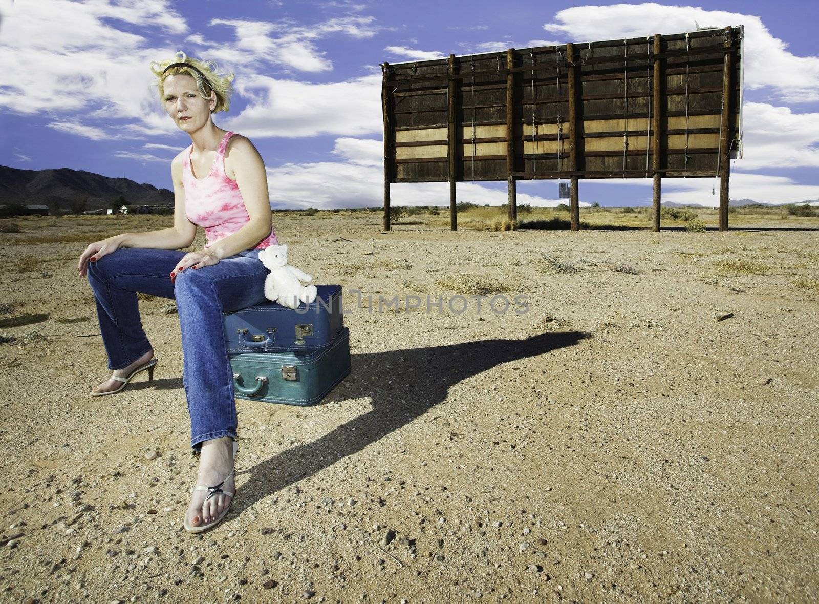 Woman sitting on suitcases in front of an old billboard waiting in the desert.