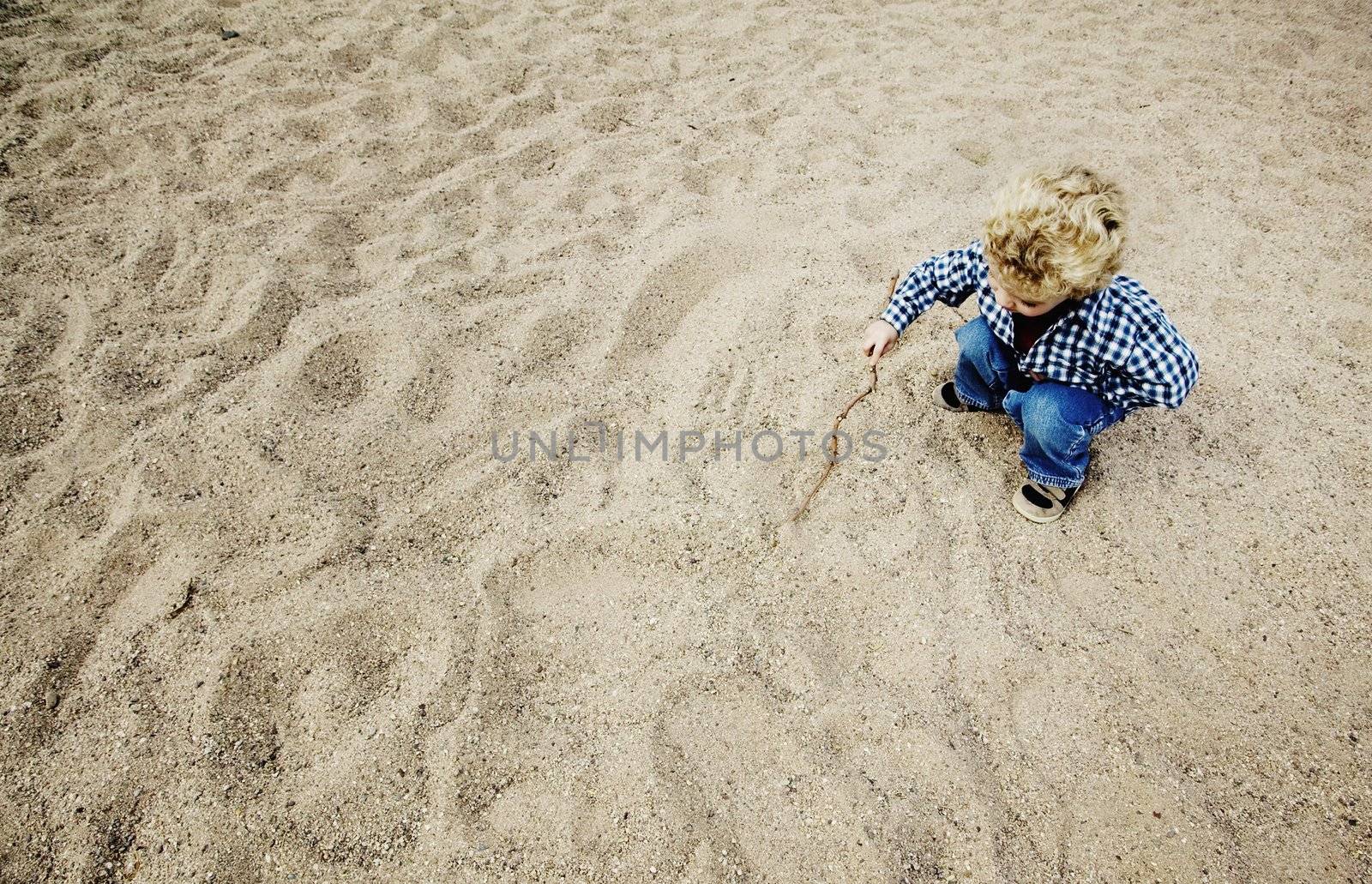 Little boy with a stick plays in a large patch of rough sand.