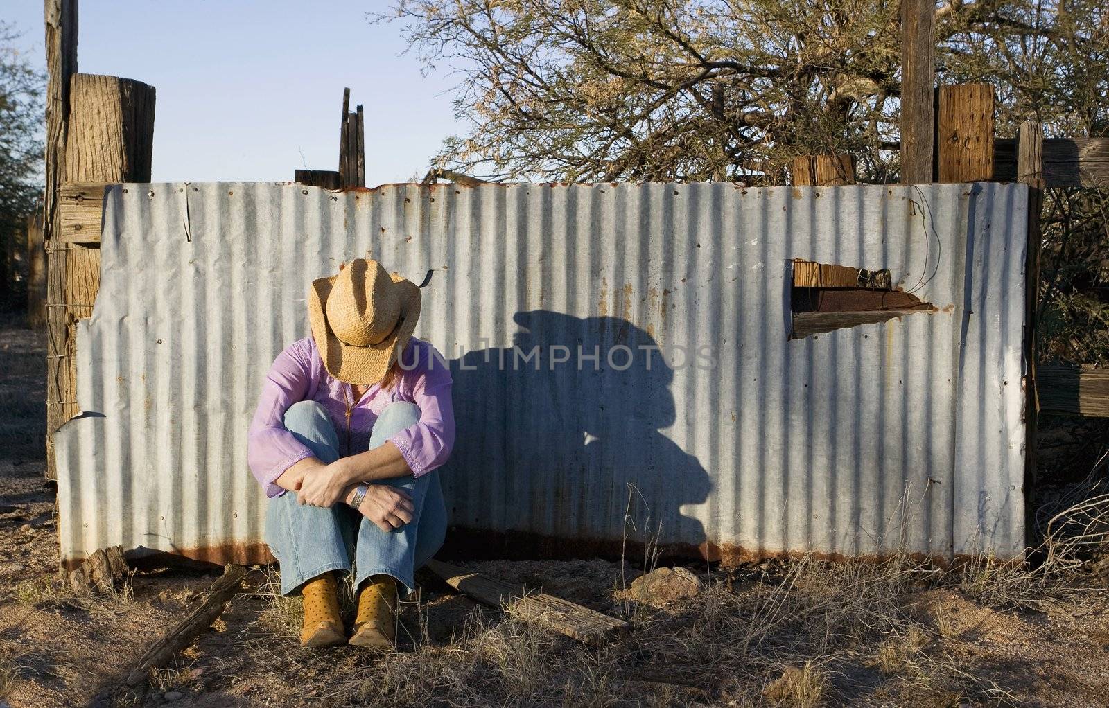 Woman wearing a straw cowboy hat with her head down leaning against a corrugated metal fence.